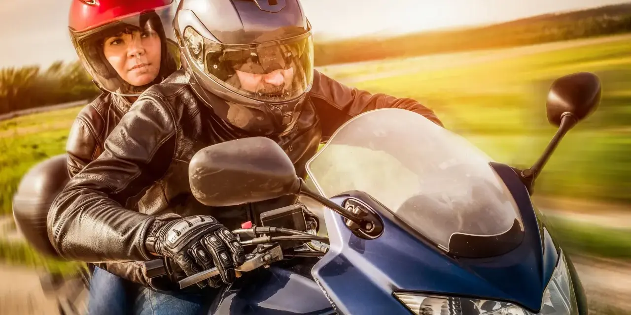 Coppia in moto, con lui alla guida e lei dietro, entrambi con caschi e giacche di pelle. Un'immagine che evoca la libertà di come guidare una moto.