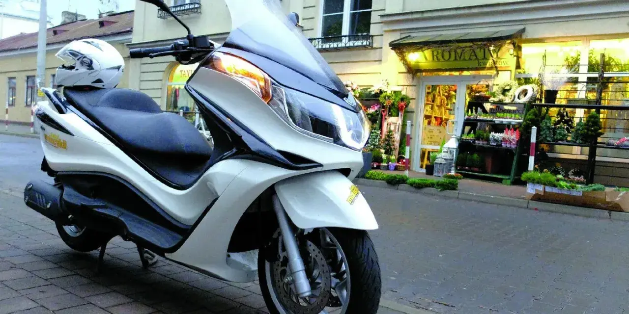 Biały skuter Piaggio X10 125, z kaskiem na siedzeniu, zaparkowany przed kwiaciarnią. Dane techniczne tego modelu są imponujące.