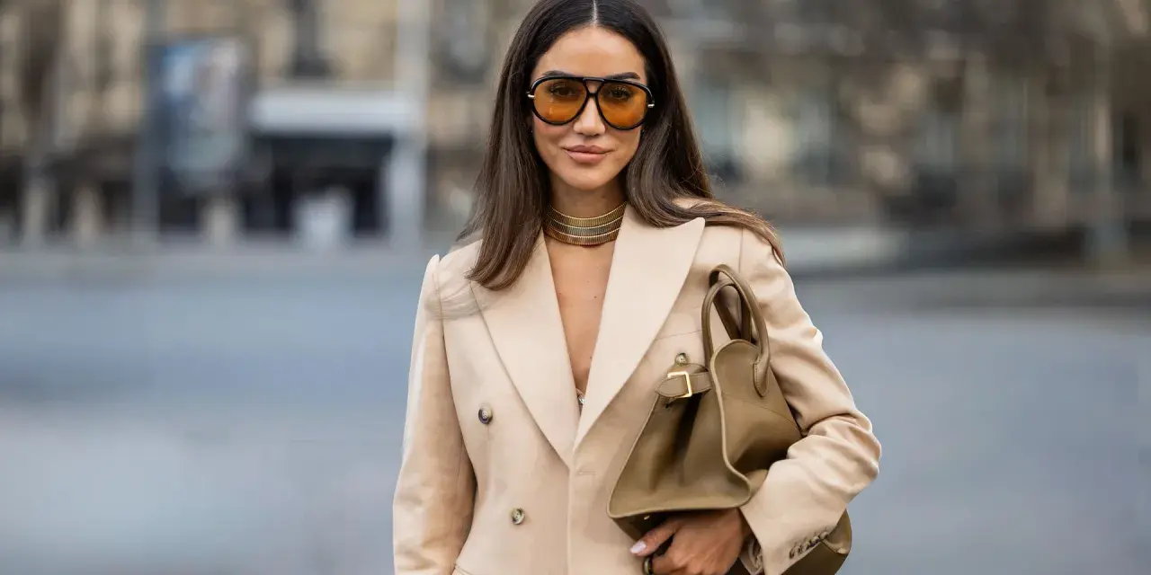 Mujer con gafas naranjas y blazer camel. Su bolso marrón y gargantilla dorada complementan el look, creando un estilo chic para combinar camel y azul.