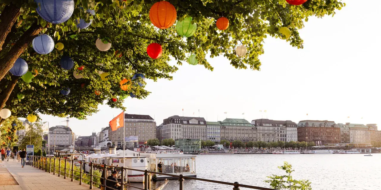Bunte Lampions schmücken einen Baum am Wasser. Im Hintergrund die elegante Skyline von Hamburg mit Gebäuden und Booten.
