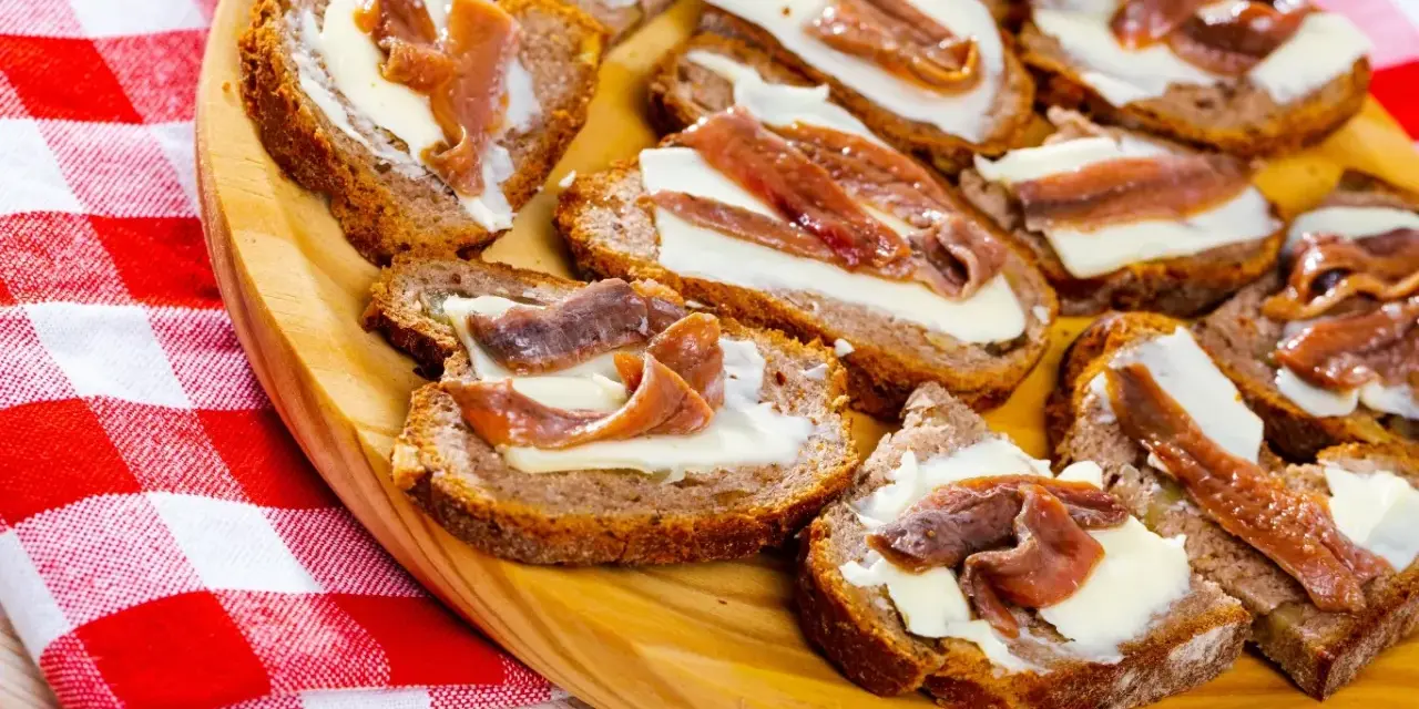 Na drewnianej desce ułożone są kromki chleba z masłem i anchois. To pyszne, proste przekąski.