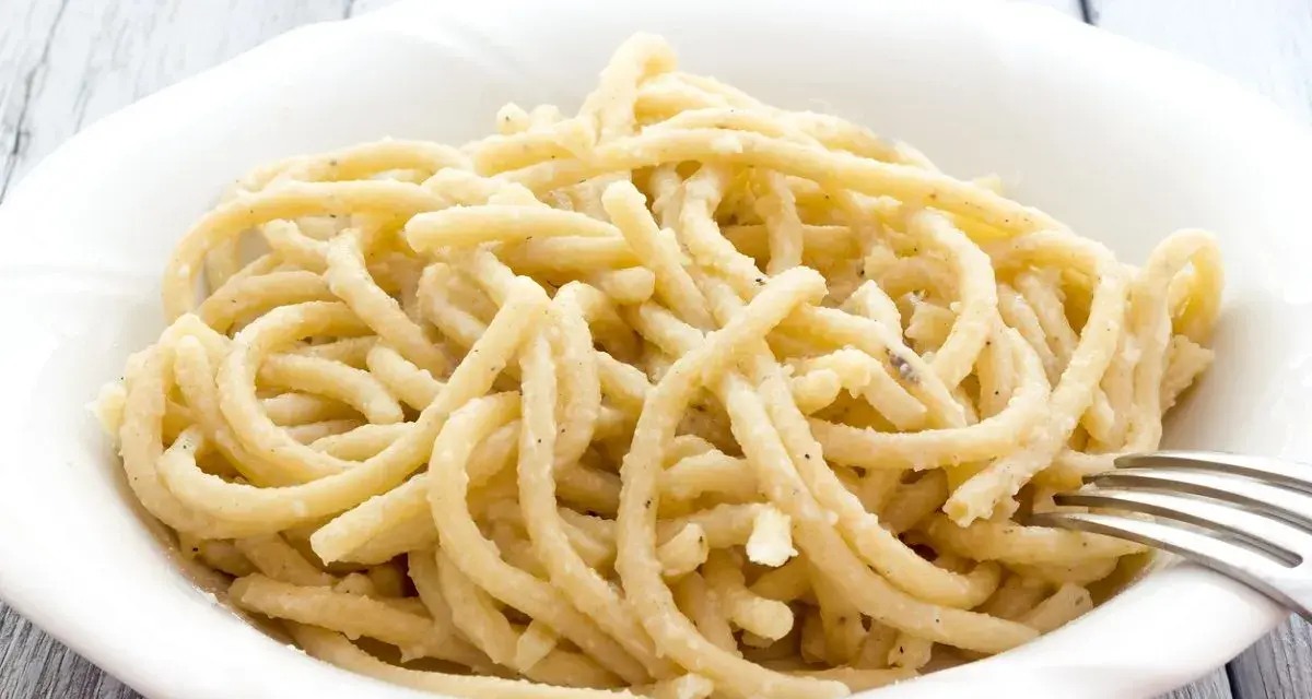 Pici, pasta fatta a mano con un'origine antica, servita in una ciotola bianca con una forchetta.
