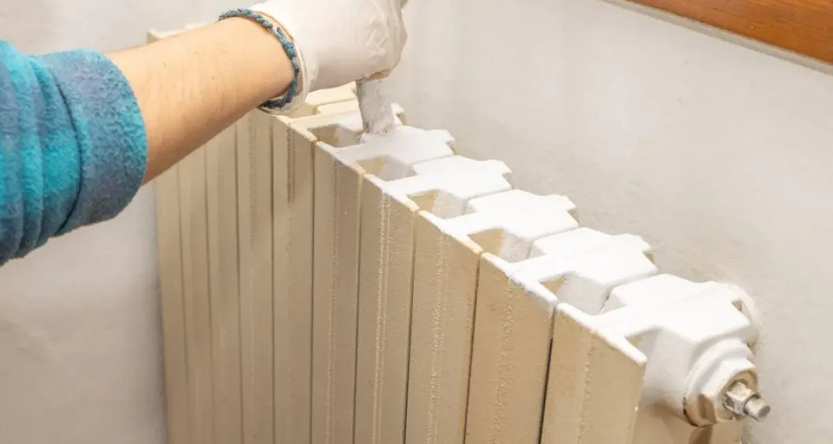 Mano con guanto bianco che dipinge un termosifone beige con vernice bianca, pronto per imbiancare la stanza.