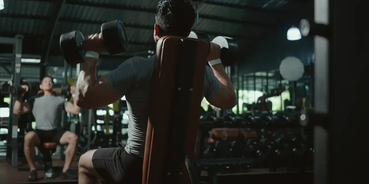 Två män tränar axlar med hantlar på ett gym. En fokuserar på push, den andra på pull.
