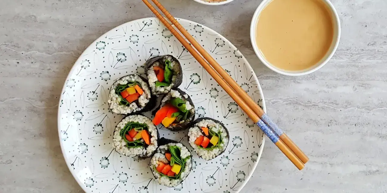 Sześć wegetariańskich rolek sushi z kolorowymi warzywami, przygotowanych z użyciem glonu do sushi, leży na talerzu obok sosów i pałeczek.