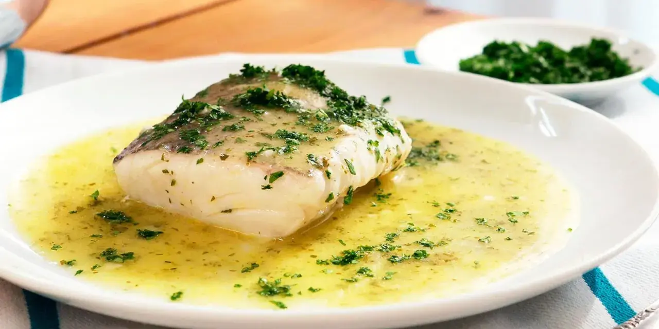 Un filete de merluza en salsa verde, con perejil picado, servido en un plato blanco.
