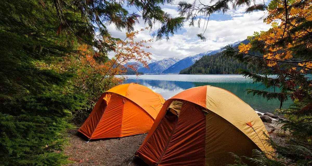 Två orangea tält står uppställda vid en sjö, omgivna av höstfärgad skog och berg. En perfekt plats för kamlunge camping.