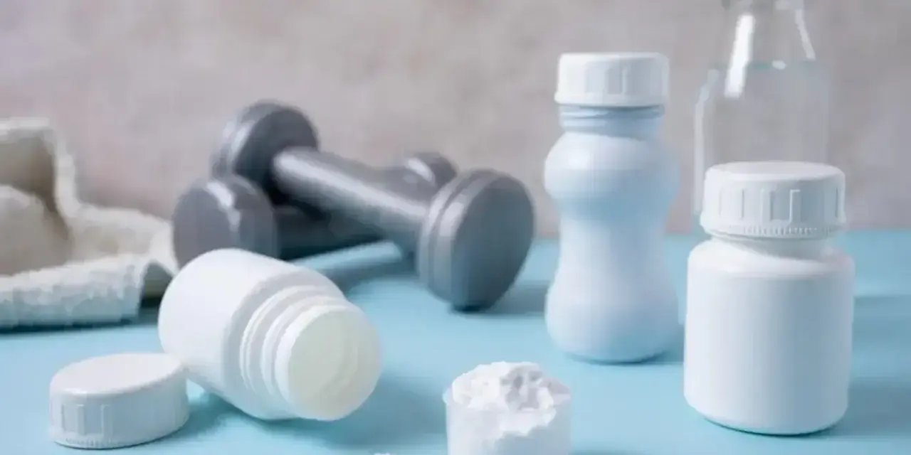 Białe pojemniki, miarka z proszkiem i hantle sugerują suplementację. Proteiny co to? To budulec mięśni, ważny dla sportowców.