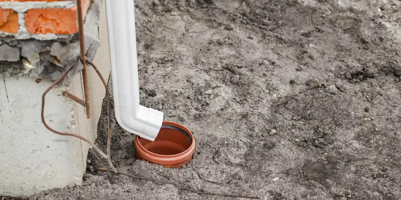 Biała rura odprowadza wodę z rynien do brązowej studzienki. Budynek z cegły w tle.