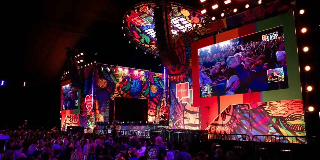 Kolorowa scena festiwalu Poland Rock 2026 z wielkim ekranem pokazującym publiczność. Tłum widzów wpatrzony w widowisko.