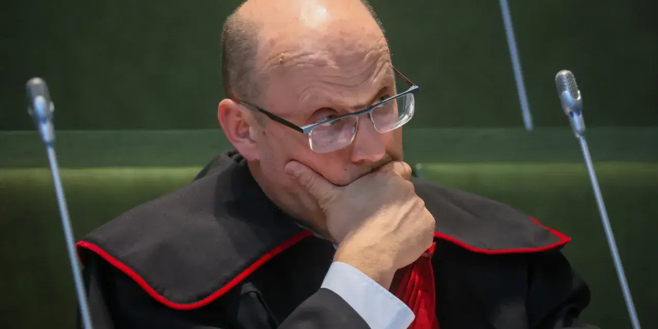 Sędzia w todze z czerwoną lamówką, w okularach, z ręką na brodzie, zamyślony.