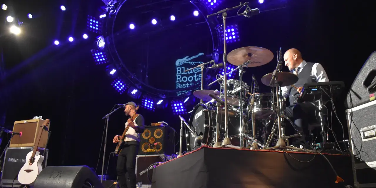 Un groupe joue du blues musique sous un grand cercle lumineux. Un bassiste à gauche, un batteur à droite.