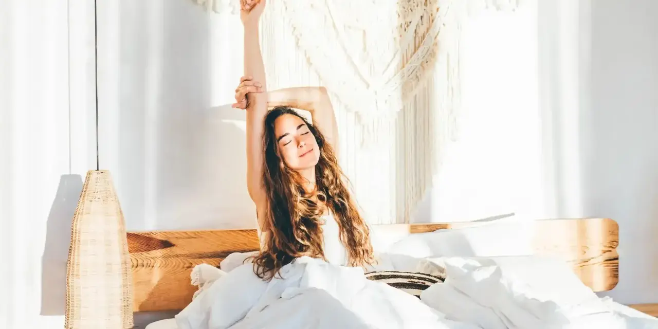Eine Frau streckt sich im Bett aus, umgeben von weißer Bettwäsche. Sie genießt den Morgen, bereit, zusammen einzuschlafen und den Tag zu beginnen.