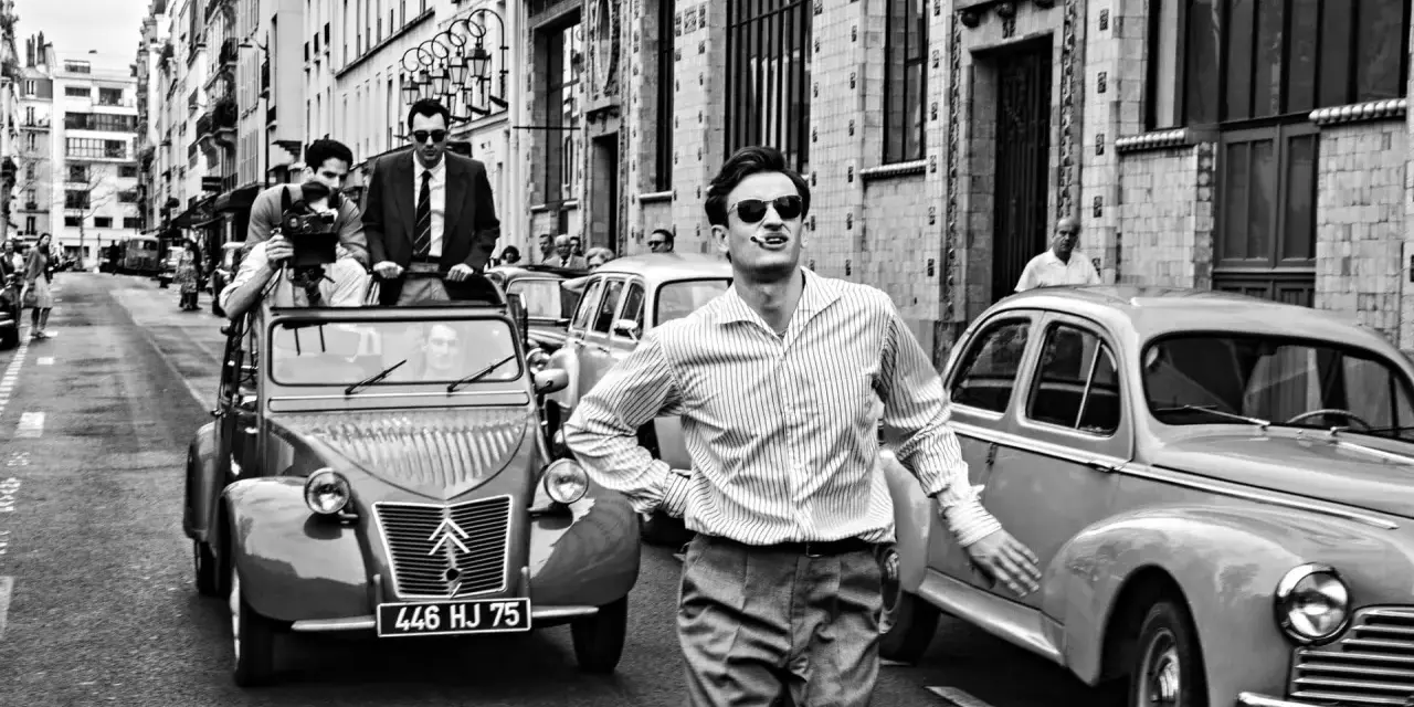 Un uomo con sigaretta in bocca, occhiali da sole e camicia a righe, in strada con auto d'epoca, evoca lo spirito de **la nouvelle vague**.