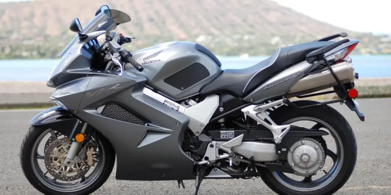 Szary motocykl Honda VFR 800 z widokiem na wodę i wzgórze w tle.