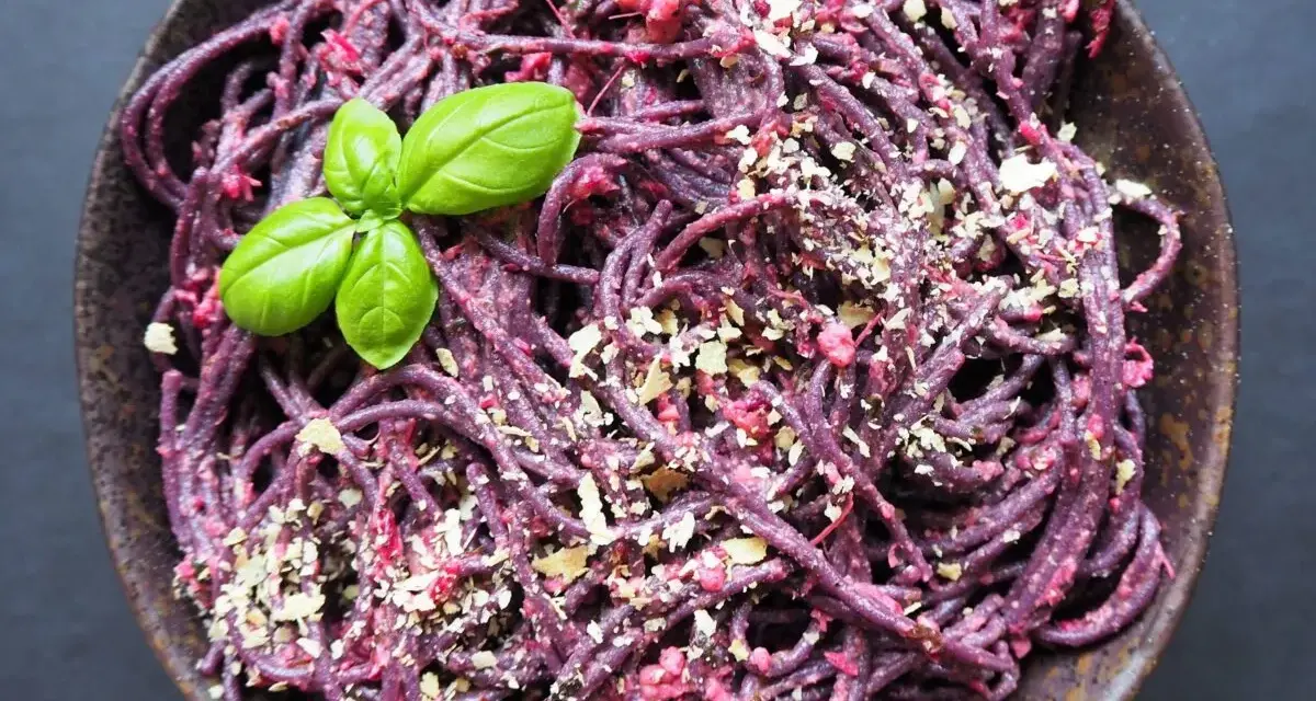 Fioletowy makaron spaghetti z buraczanym pesto, posypany orzechami i udekorowany listkiem bazylii.