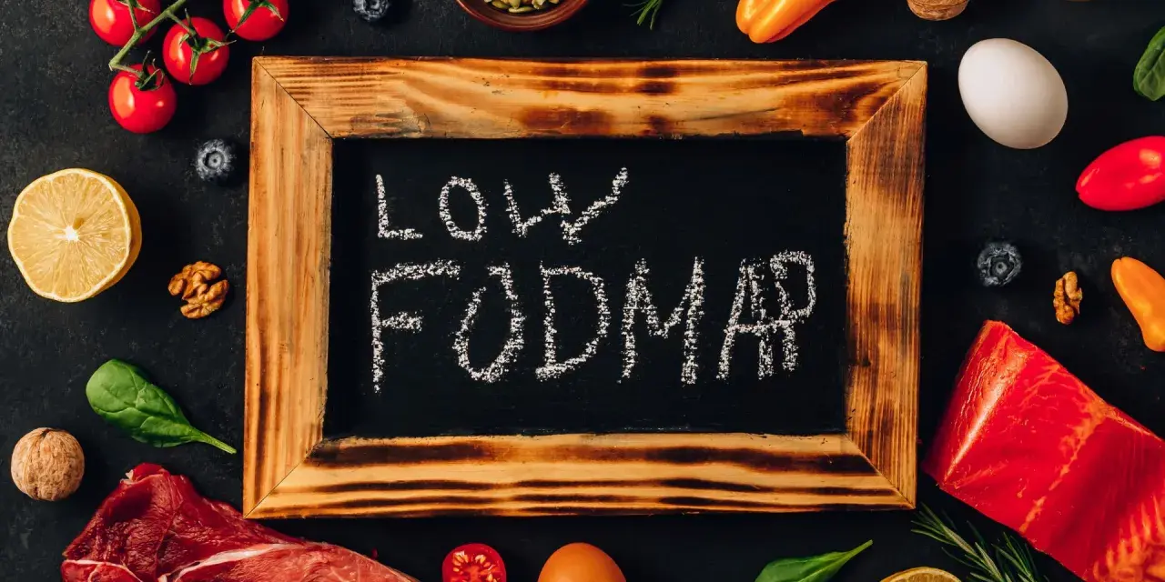 Na tablicy napis "LOW FODMAP". Wokół świeże produkty: mięso, ryba, pomidorki, cytryna, jajko, papryka, orzechy, jagody, szpinak.