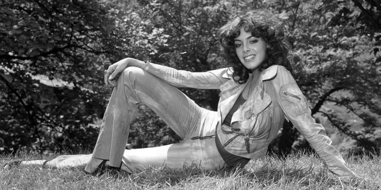 Nadia Cassini, con il suo fisico prorompente, posa su un prato, sfoggiando un completo giacca e pantaloni.