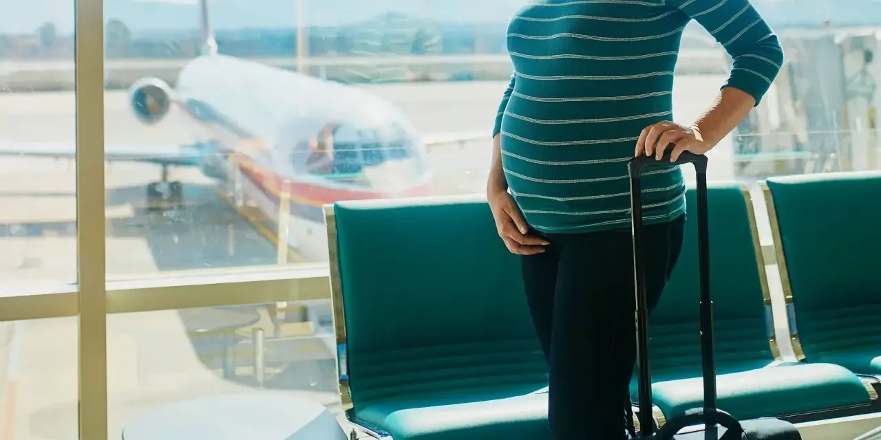 Ciężarna kobieta z walizką czeka na lotnisku. W tle widać samolot i pasażerskie fotele. Latanie samolotem w ciąży to wyzwanie, ale podróż dobiega końca.