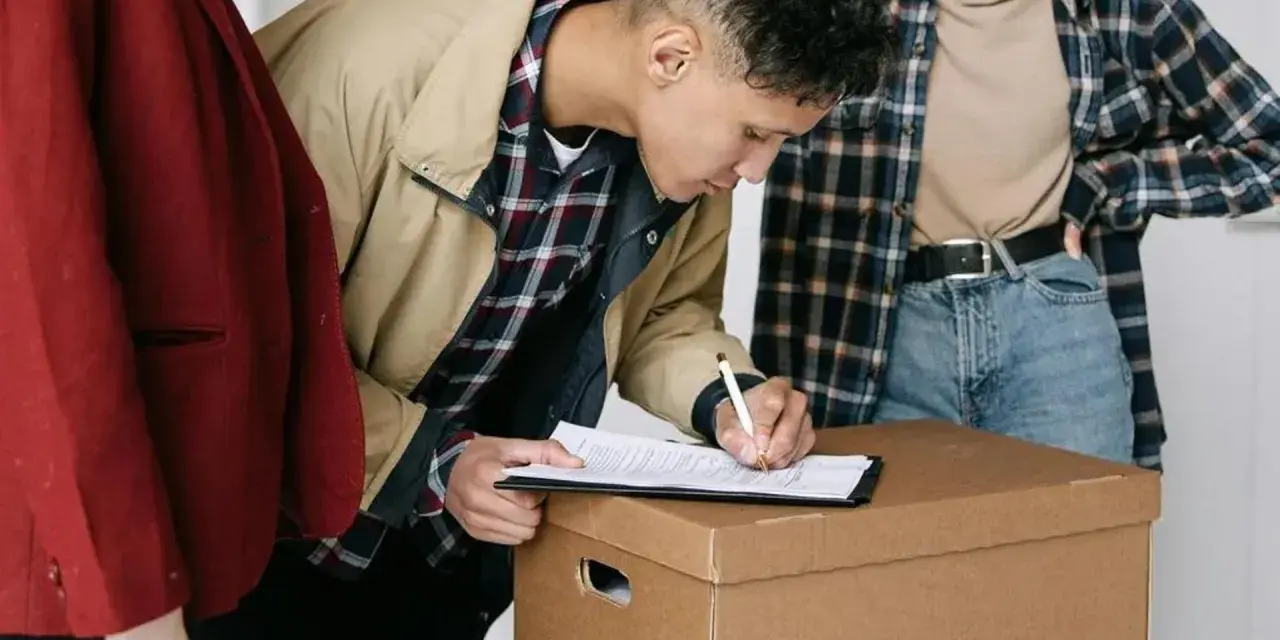 Joven firma contratos de alquiler sobre una caja de mudanza, acompañado de dos personas.