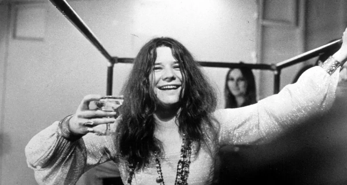 Janis Joplin z kieliszkiem w dłoni, uśmiechnięta. Jej charakterystyczne włosy i naszyjniki przywodzą na myśl jej niezapomniane utwory.