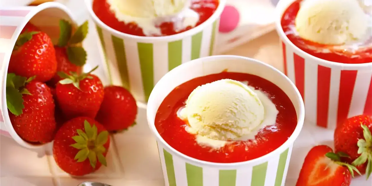 Vanilleeis mit Erdbeeren und Sahne in bunten Bechern. Frische Erdbeeren liegen daneben.