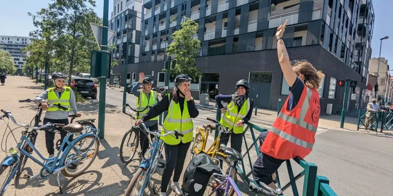 Des adultes souriants, casqués et en gilets fluo, prêts à apprendre à faire du vélo adulte. Ils sont avec leurs vélos, devant un immeuble moderne.