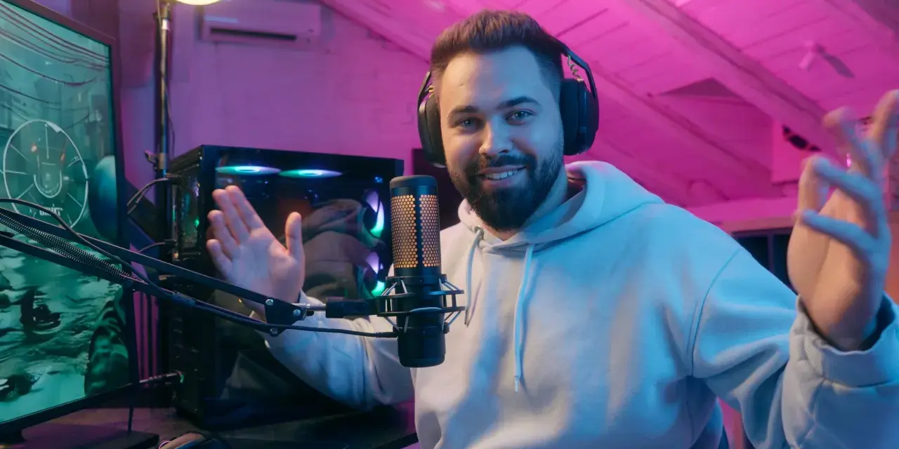 Uśmiechnięty streamer w słuchawkach, przy mikrofonie, tłumaczy, streaming co to jest. W tle komputer i różowe oświetlenie.