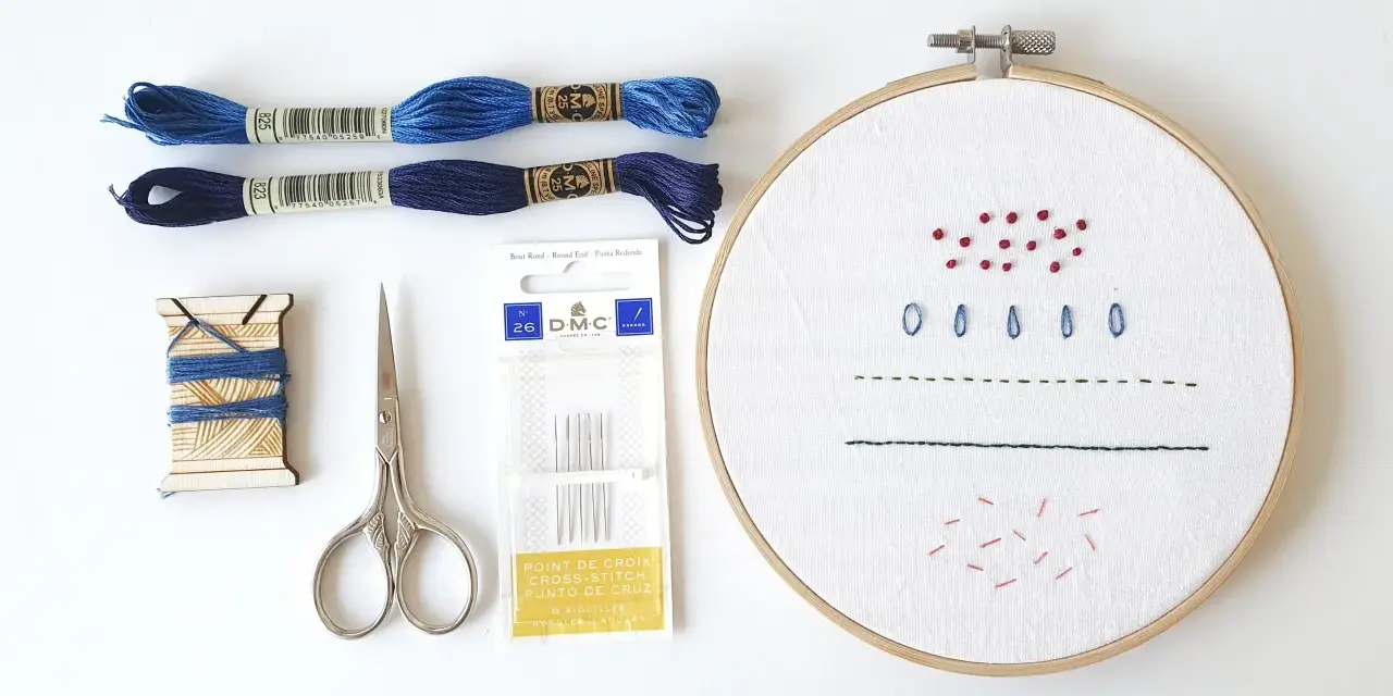Materiali per un tutorial ricamo: fili blu, forbici, aghi e tela con primi punti.