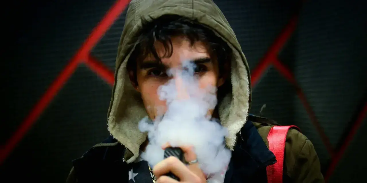 Joven con capucha exhala vapor de un vapeador, con un diseño de estrellas en su camiseta. Los nicokits son su secreto.