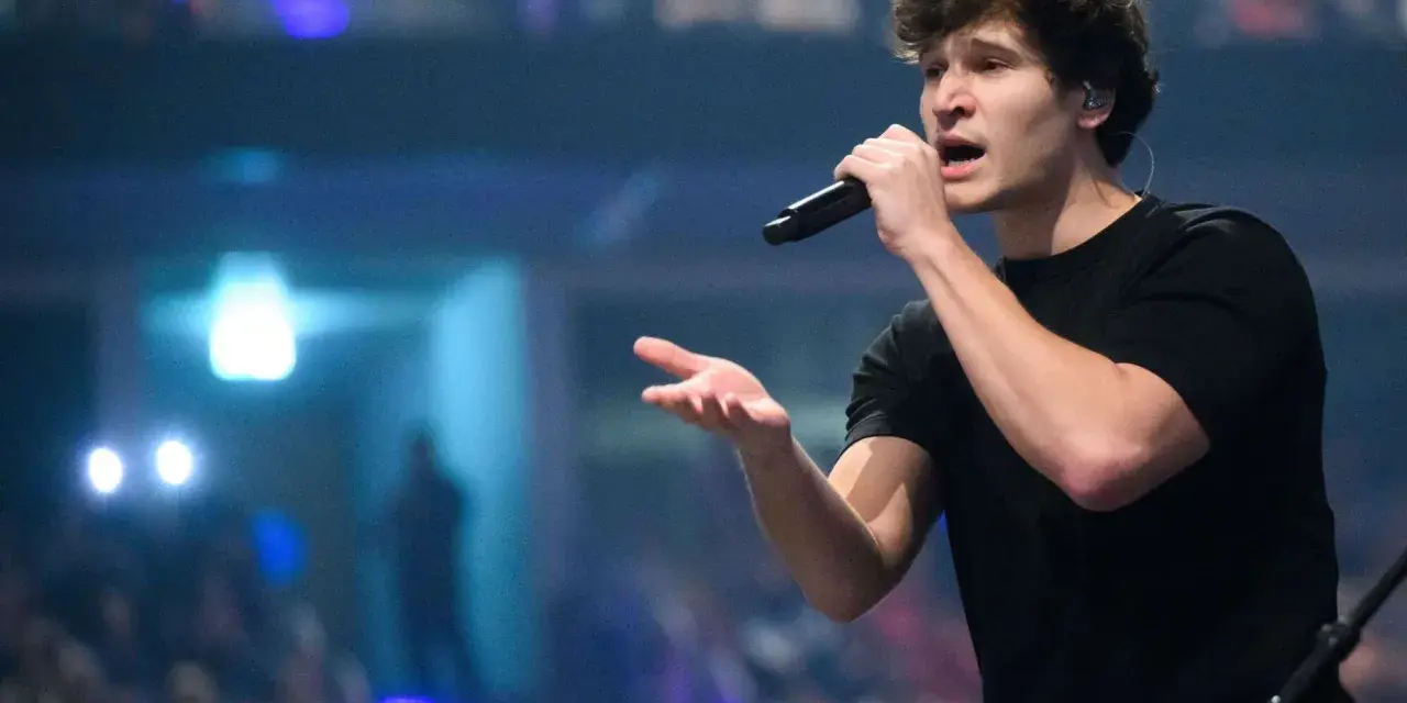Ein junger Mann mit Mikrofon singt auf einer Bühne, im Hintergrund blaue Lichter.