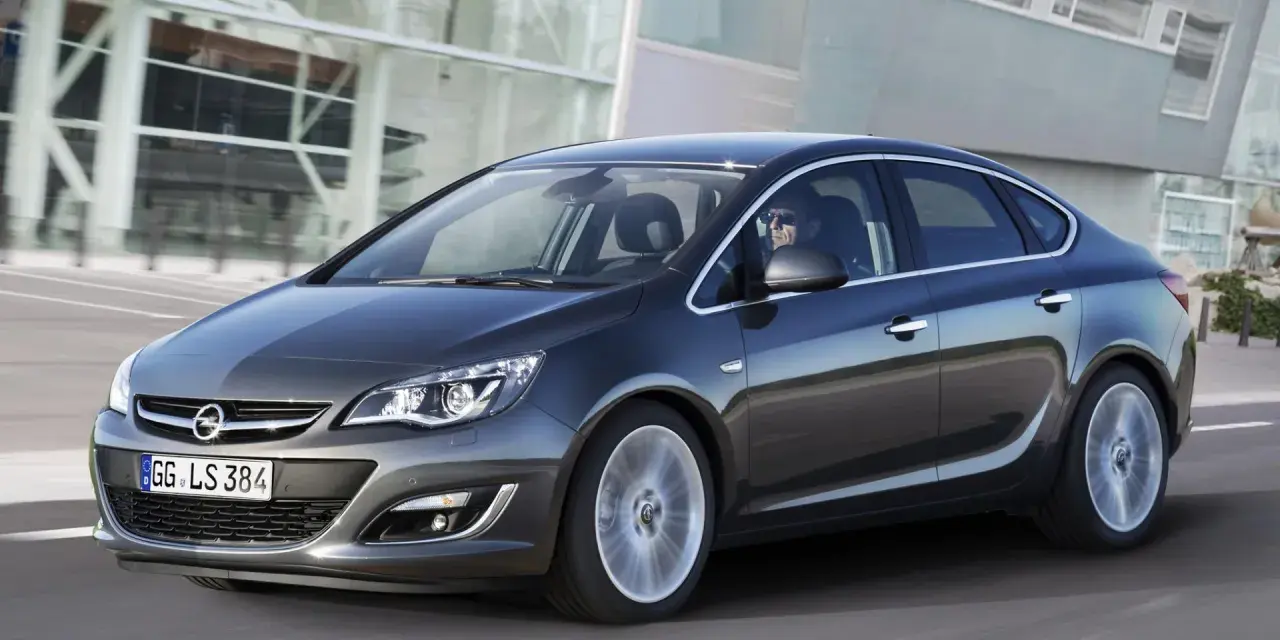 Ciemnoszary Opel Astra J 1.6 benzyna w ruchu, z kierowcą w okularach za kierownicą, na tle nowoczesnego budynku.