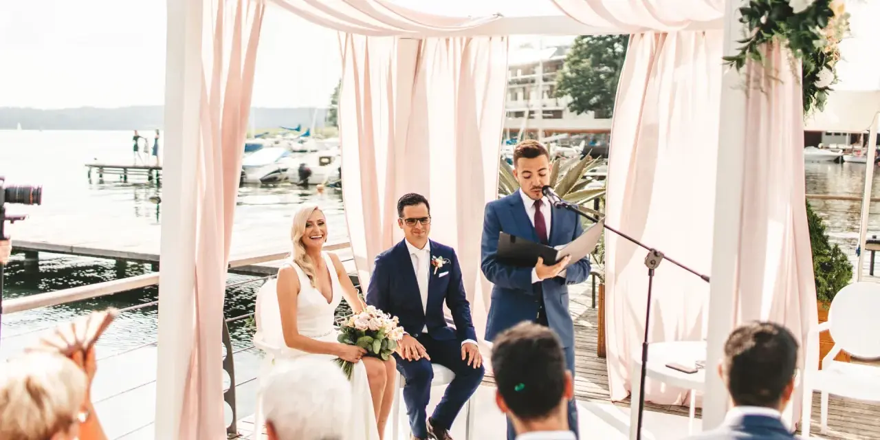 Ślub humanistyczny co to? Para młoda, urzędnik i goście nad jeziorem. Ceremonia w białej altanie z różowymi zasłonami.
