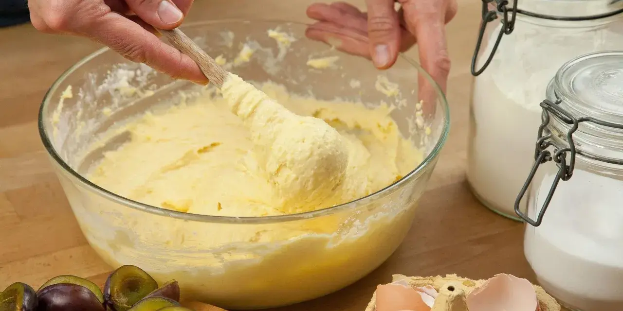 Hände rühren Teig mit Butter oder Margarine in einer Schüssel. Daneben liegen Pflaumen, Eier und Zucker.