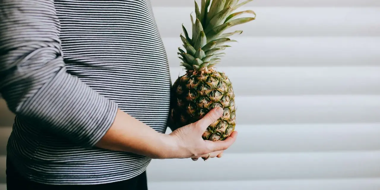 Brzuch kobiety w paski, trzymająca ananasa. Wygląda jak ananas w ciąży, symbolizujący zdrową dietę.