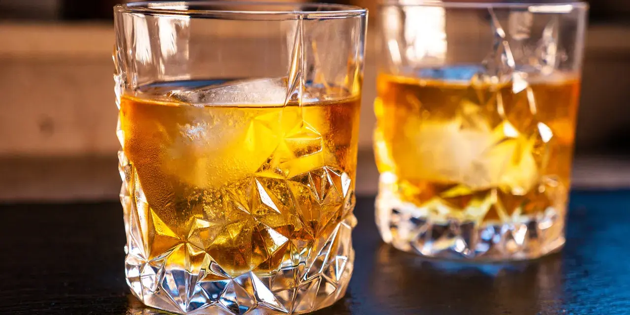 Zwei Gläser mit bernsteinfarbenem on the rocks drink, beleuchtet von warmem Licht.