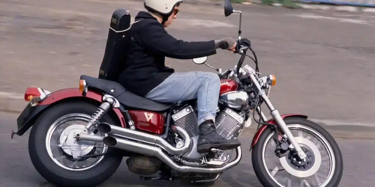 Mężczyzna w kasku jedzie na czerwonym motocyklu Yamaha Virago 250 z futerałem na plecach.