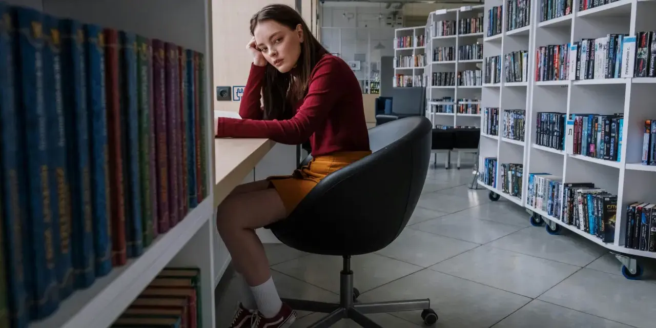 Zamyślona nastolatka w bibliotece, otoczona książkami, odzwierciedla złożoność zachowań nastolatków w okresie dojrzewania.