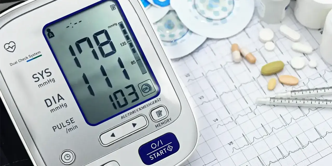 Cyfrowy ciśnieniomierz pokazuje wysokie ciśnienie 178/103. Obok leżą tabletki, elektrody EKG i papier z zapisem EKG, sugerując monitorowanie holterem ciśnieniowym.