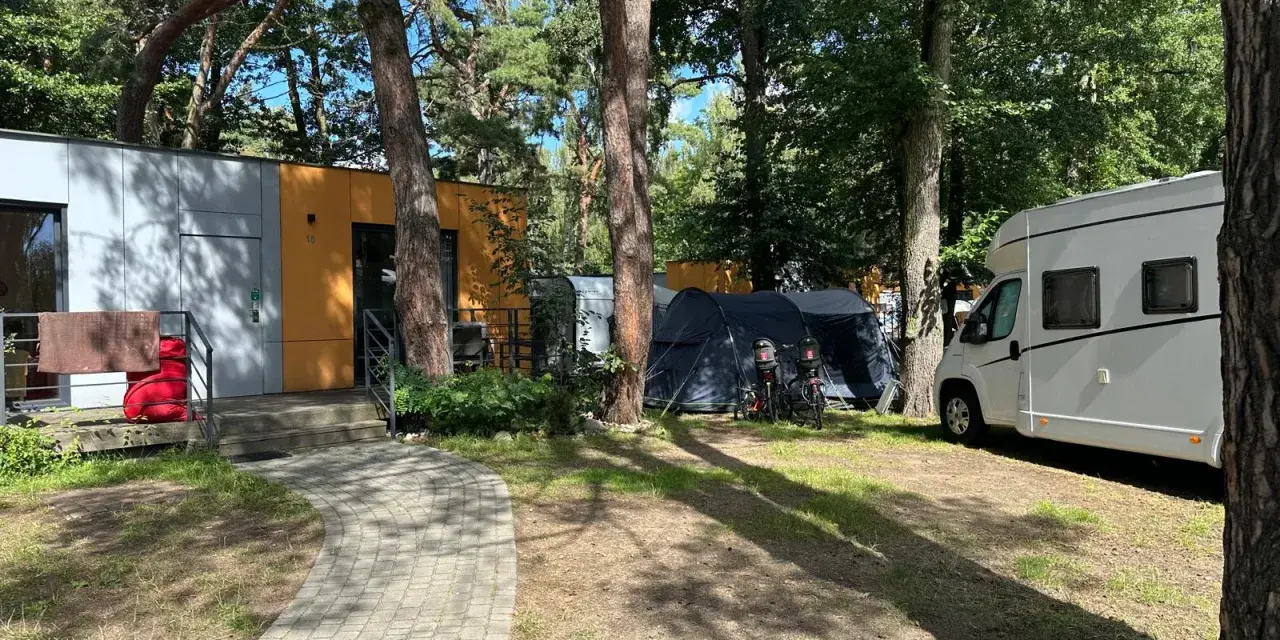 Na kempingu 45, wśród drzew, stoją nowoczesne domki, namioty i biała kamper.