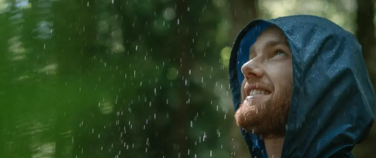 Mann mit Bart blickt lächelnd in den Regen, seine blaue Regenjacke hält ihn trocken.