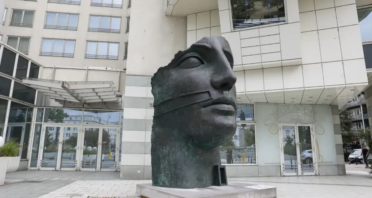 Okaleczona, ciemna rzeźba głowy z opaską na ustach, na tle nowoczesnego budynku.