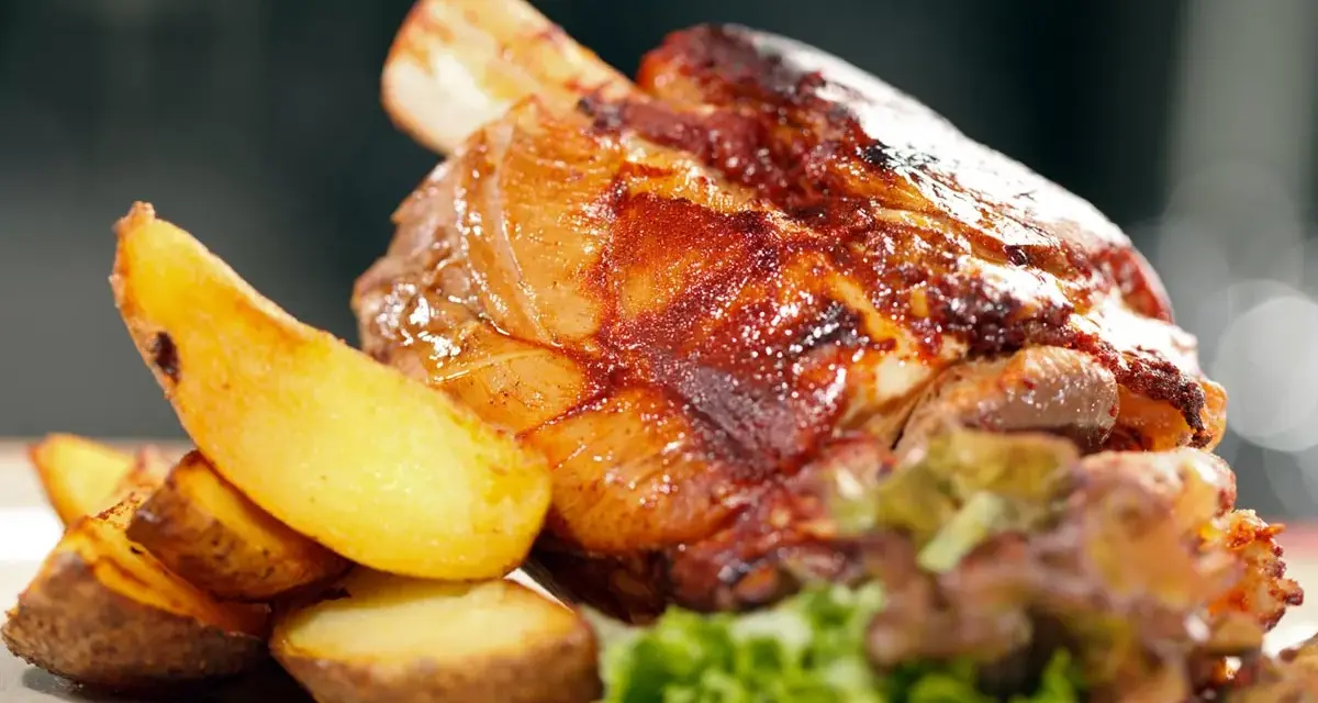 Codillo de cerdo asado, crujiente y jugoso, acompañado de patatas gajo doradas y una guarnición verde.