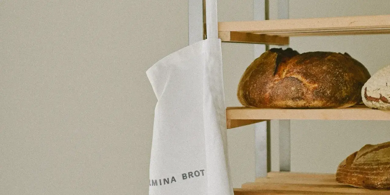 Ein weißer Stoffbeutel mit der Aufschrift "WILMINA BROT" hängt neben einem Regal mit frisch gebackenen Broten.
