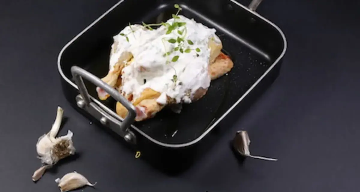 Un poulet entier est nappé de fromage blanc crémeux et garni d'herbes fraîches dans un plat noir. Des gousses d'ail sont disposées autour.