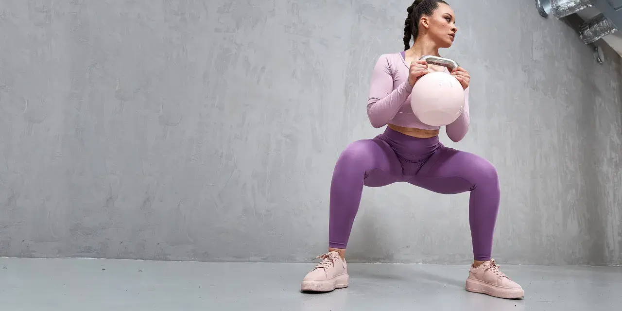 En kvinna i lila träningskläder gör en kettlebell swing-rörelse, med en ljusrosa kettlebell i händerna.