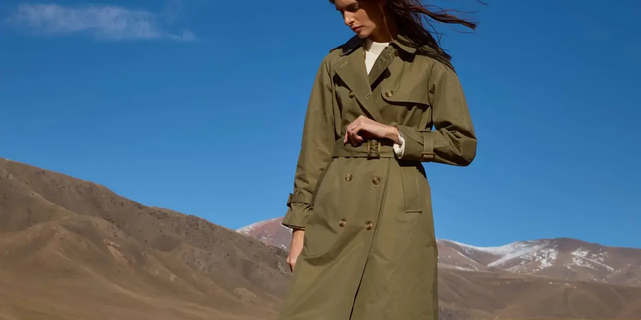 Mujer con un elegante trench color caqui, con el pelo al viento, en un paisaje montañoso bajo un cielo azul.