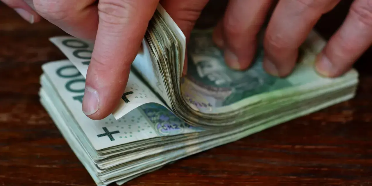 Dłonie przewracają stos banknotów 100 zł na drewnianym blacie.
