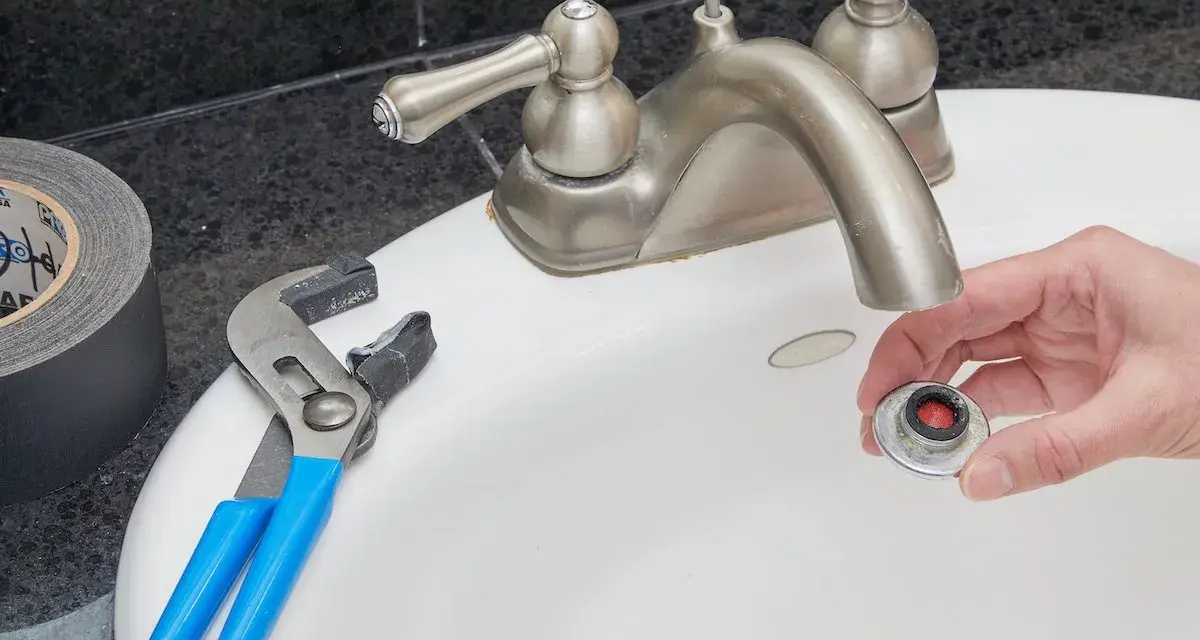 Hand holding a removed faucet aerator with red buildup, next to pliers and duct tape on a sink.