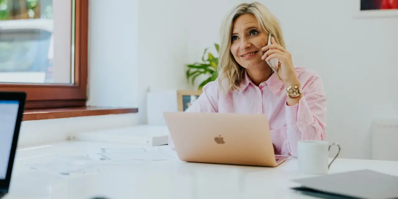Kobieta w różowej koszuli rozmawia przez telefon, siedząc przy biurku z laptopem.